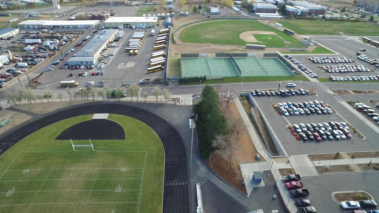 Opening aerial frame over Hidden Lake, Colorado, showing a black running track and football field below, tennis courts and parked cars in the middle distance, baseball fields beyond, and no visible people.