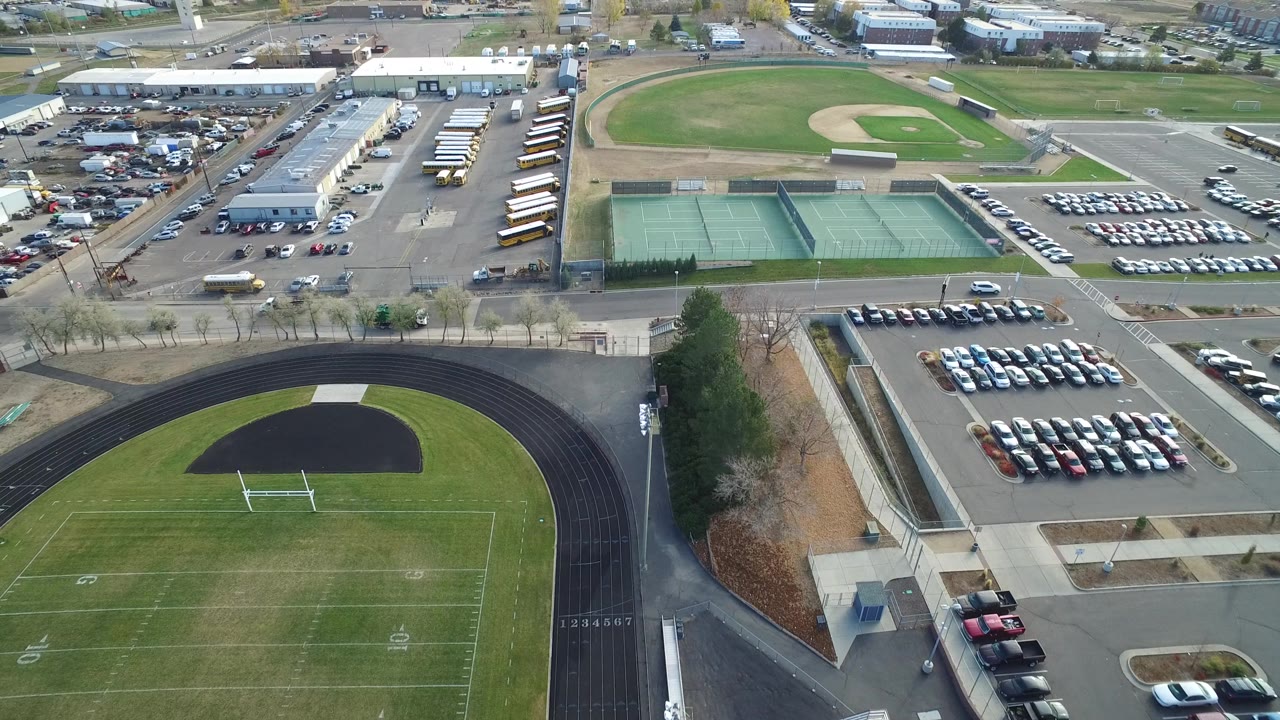 Aerial frame in Hidden Lake, Colorado, with the curved track and field lower in view, green tennis courts centered left, rows of parked cars to the right, buses near low buildings, and no visible people.