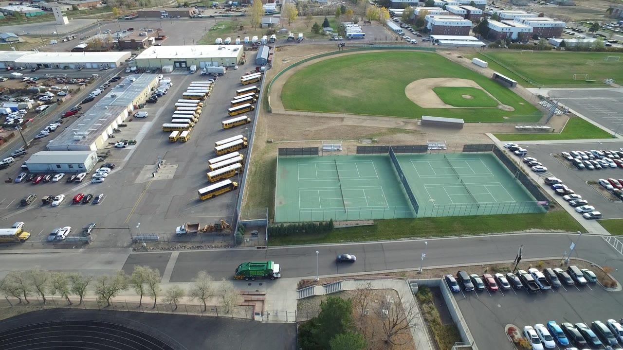 At one second, the aerial view in Hidden Lake, Colorado centers the tennis courts and road while the baseball field, school bus rows, parking lots, and low commercial buildings sit farther back, with no visible people.