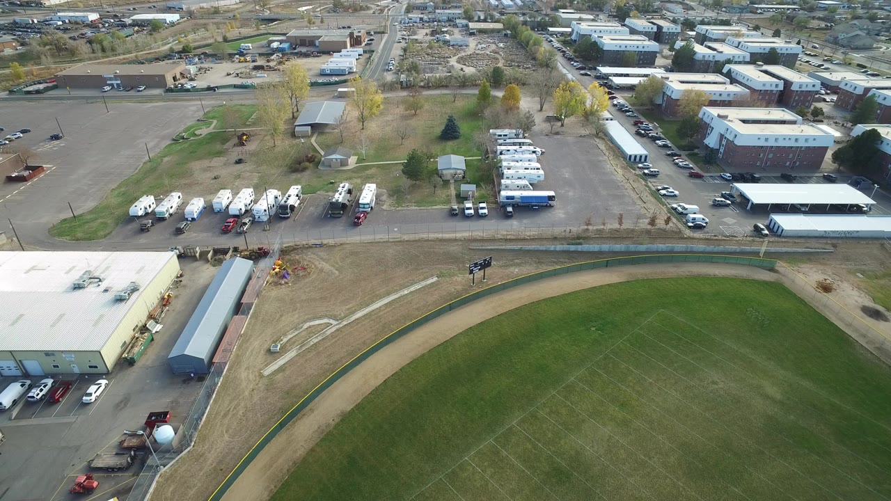 Elevated drone view in Hidden Lake, Colorado showing the edge of a green ball field below low buildings, parked vehicles, roads, trees, and no visible people.