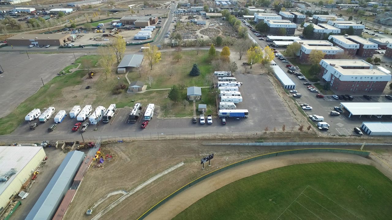 Aerial view over parking lots, trailers, low buildings, and a ball field edge.