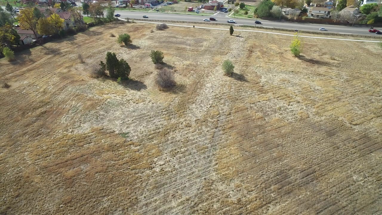 Aerial frame in Hidden Lake, Colorado showing a dry open field with scattered trees, a highway along the top edge, distant homes, and no visible people.