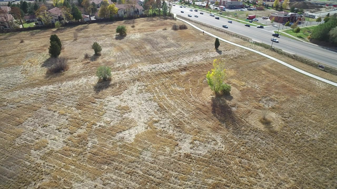 Aerial view in Hidden Lake, Colorado showing dry open grassland, scattered trees, a road with cars near the top edge, houses beyond, and no visible people.