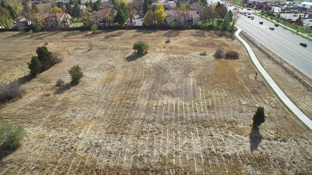 Aerial view in Hidden Lake, Colorado showing a tan plowed open field, scattered trees, a sidewalk, a multi-lane road with cars, and houses along the top edge, with no visible people.