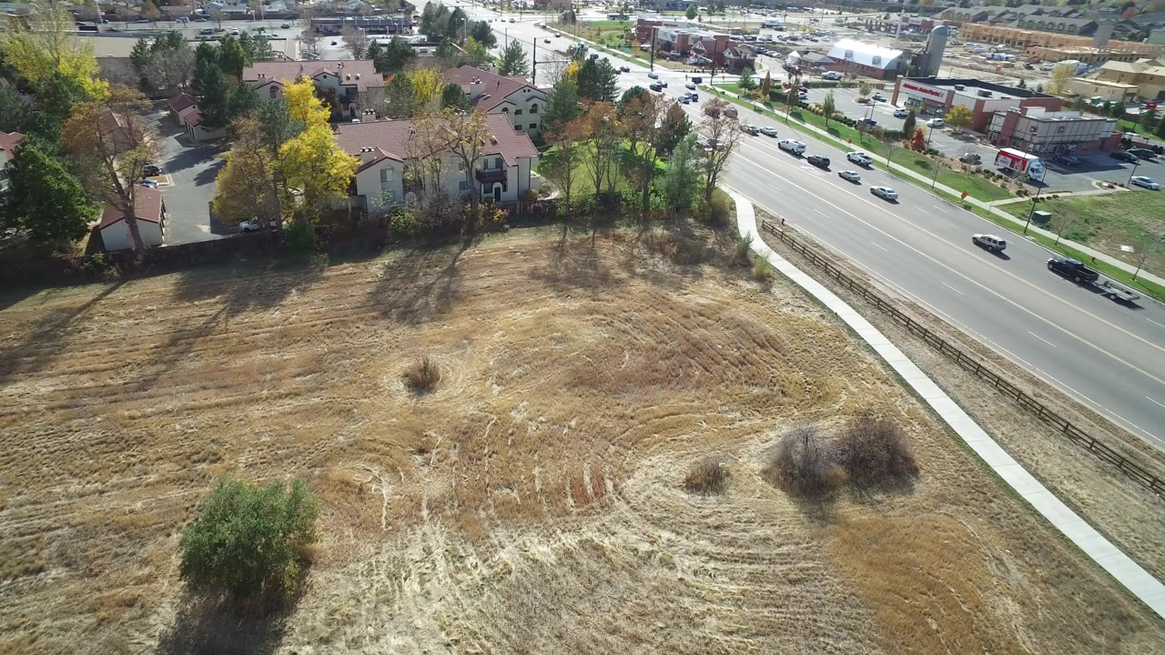 High aerial frame in Hidden Lake, Colorado showing dry open ground, a sidewalk, a multilane road with cars, red-roofed houses, trees, and no visible people.
