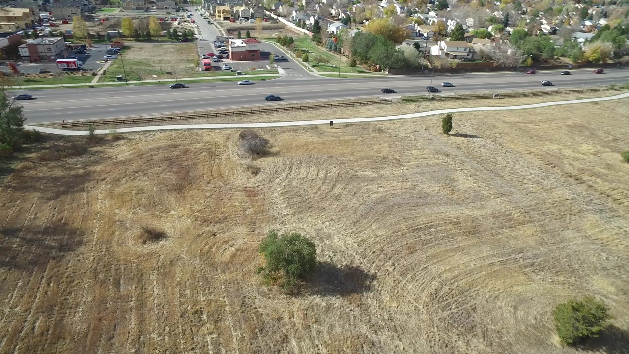 Aerial frame over Hidden Lake, Colorado, showing a dry mowed field, scattered trees, a highway with cars, and residential blocks beyond, with no visible people.