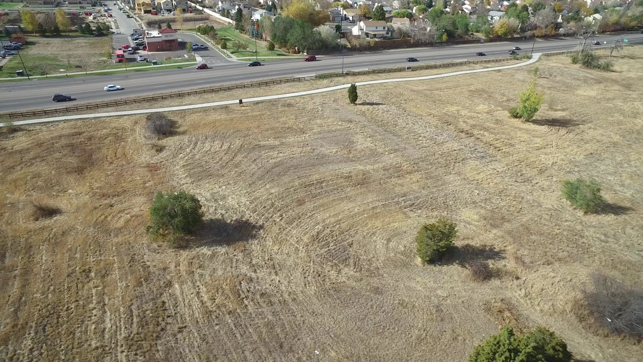 Aerial frame over Hidden Lake, Colorado showing dry plowed open ground with scattered trees, a road with cars along the top edge, distant houses, and no visible people.