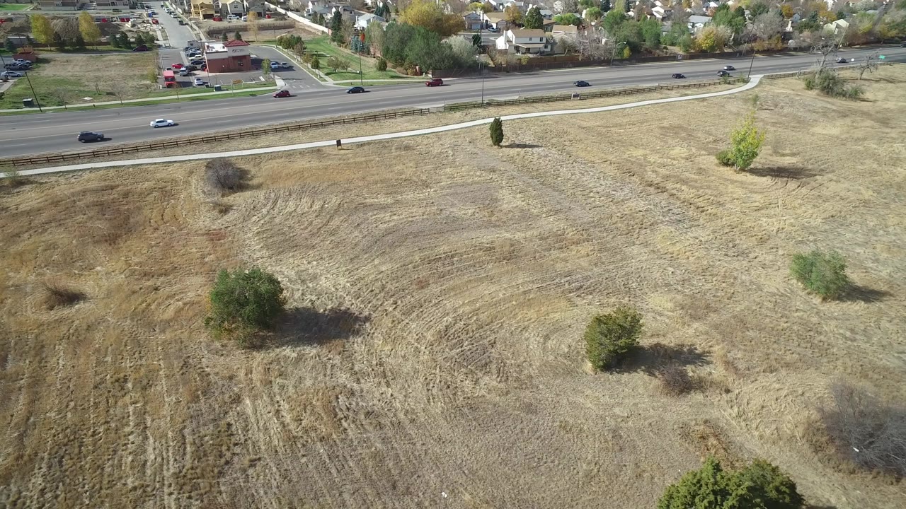 A slightly advanced aerial view in Hidden Lake, Colorado frames tan open land with curved plow marks, small trees, passing cars on the road, distant houses, and no visible people.