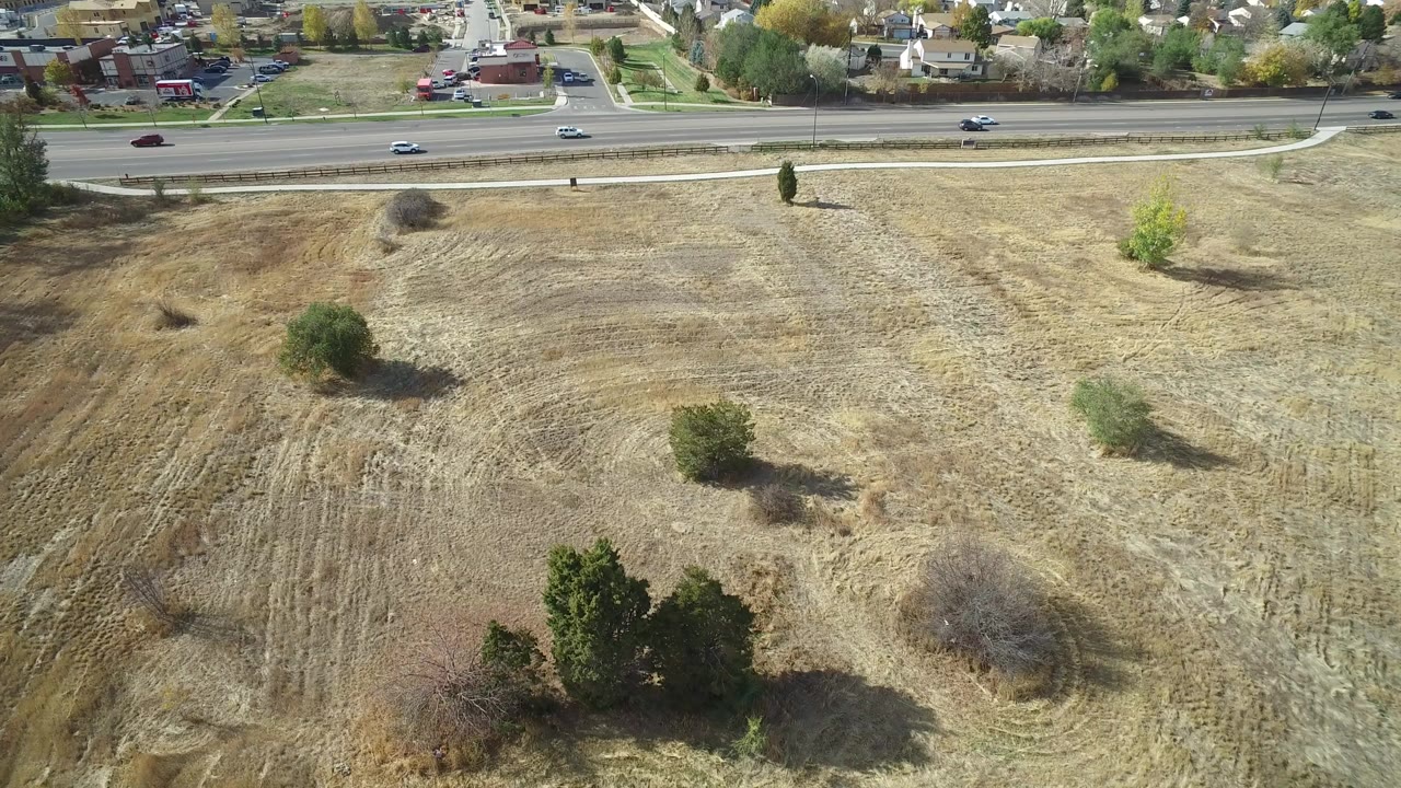 Opening aerial frame over a dry roadside field in Hidden Lake, Colorado, with scattered trees, cars on the road, homes beyond it, and no visible people.