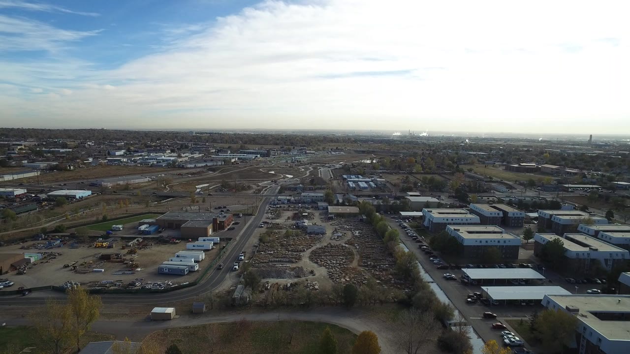 Aerial frame over Hidden Lake, Colorado, showing low rooftops in the foreground, road lanes through industrial lots, distant development, cloud cover, and no visible people.