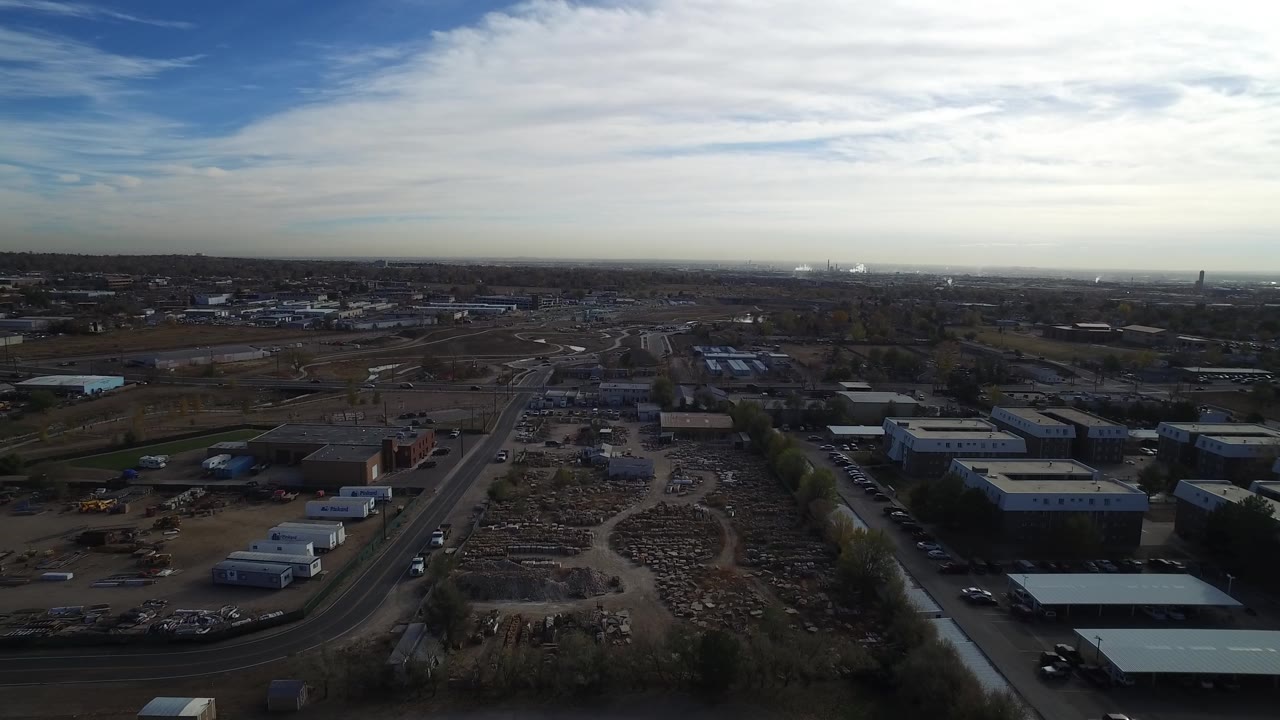 Wide aerial view in Hidden Lake, Colorado over industrial yards, warehouse roofs, curved roads, distant suburbs, and a bright cloudy sky with no visible people.
