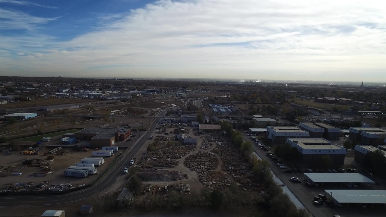 Aerial frame in Hidden Lake, Colorado looking over warehouse buildings, open material yards, parked vehicles, road lanes, and distant residential blocks with no visible people.