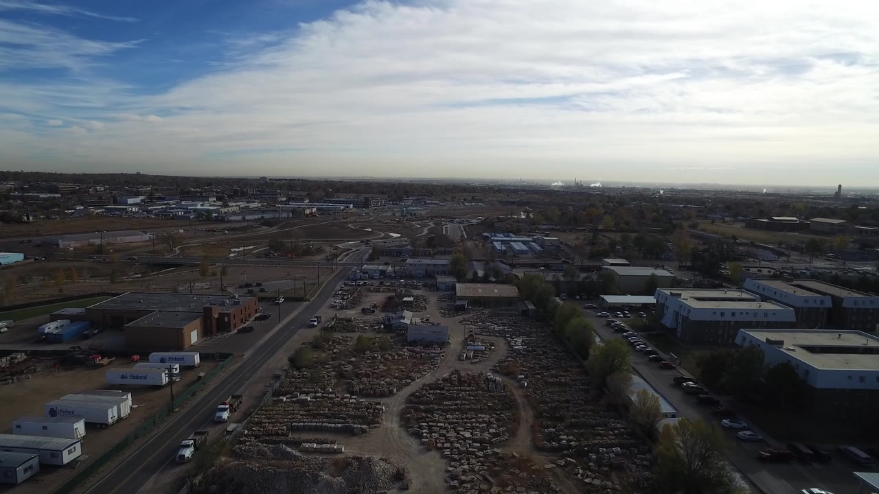 Aerial view over roads, low buildings, stacked yard materials, and a cloudy sky.