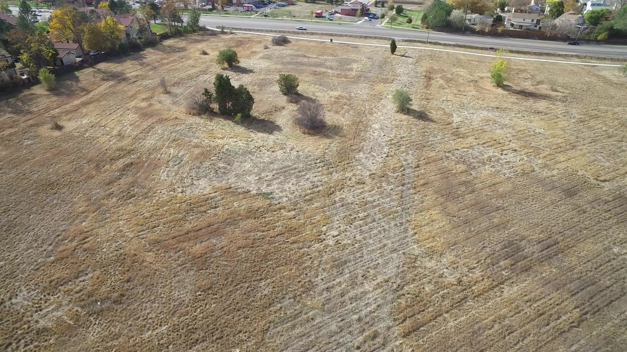 High oblique aerial frame over Hidden Lake, Colorado, showing dry brown open ground, scattered trees, a road with small cars, nearby houses, and no clearly visible people.