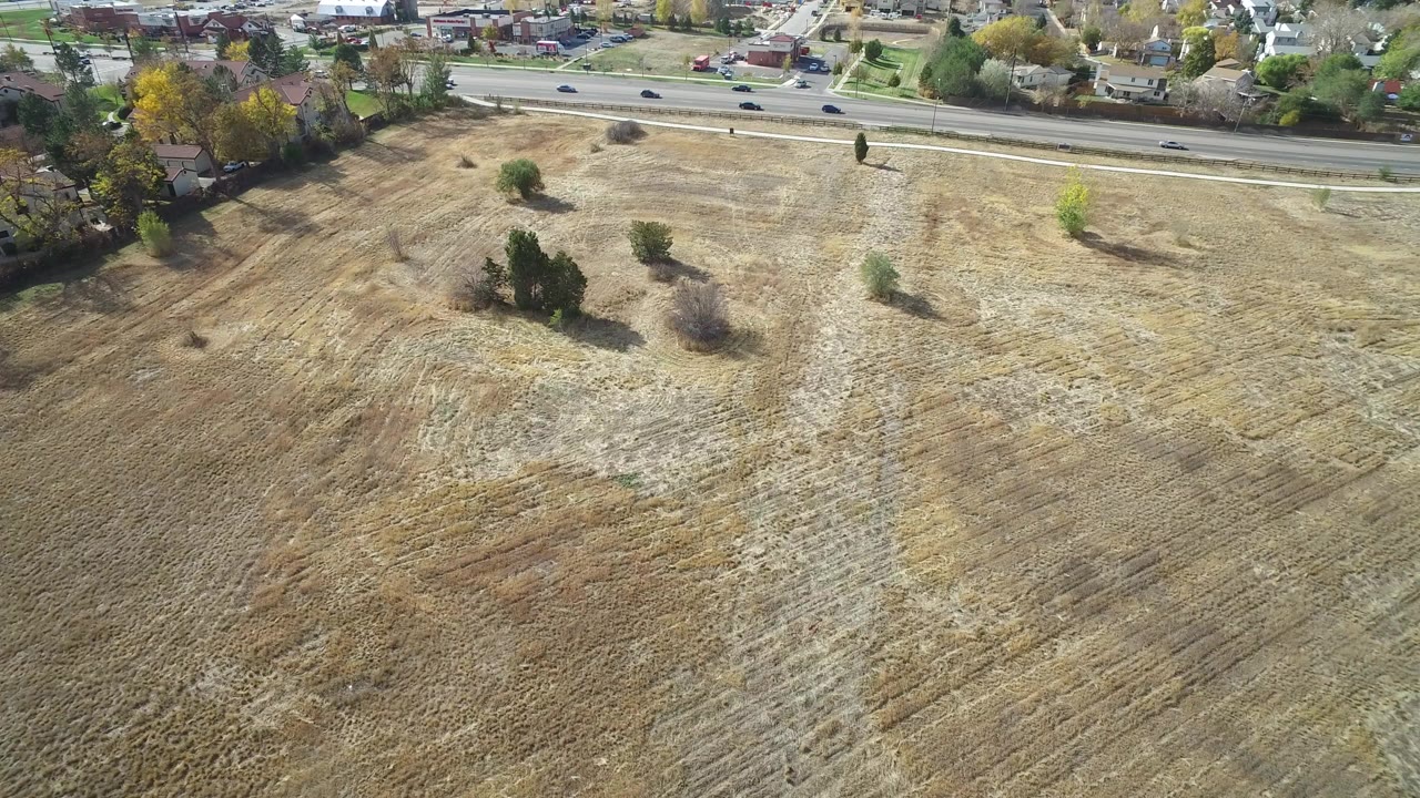 Aerial frame from Hidden Lake, Colorado showing a dry tan field with scattered trees, a paved road across the upper frame, homes beyond the road, and no visible people.