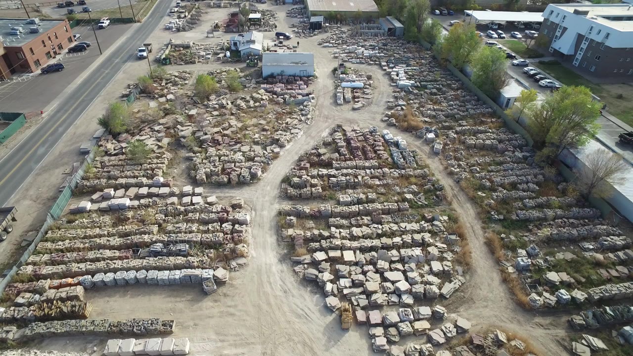 High oblique aerial view over a Hidden Lake, Colorado industrial yard with pale dirt lanes, rows of stone or concrete materials, a paved road at left, nearby buildings, and no visible people.