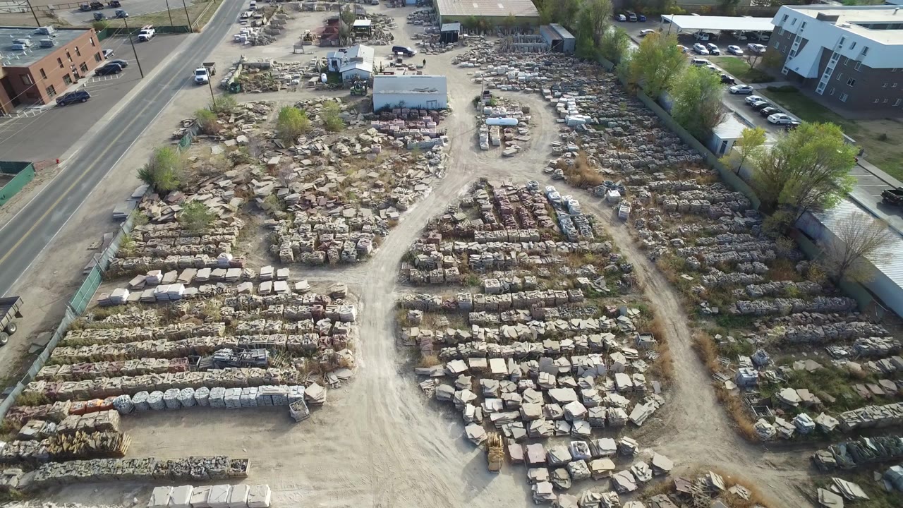 Aerial frame over Hidden Lake, Colorado showing the central dirt road, organized rows of stacked materials, parked vehicles near a small building, trees along the edge, and no visible people.