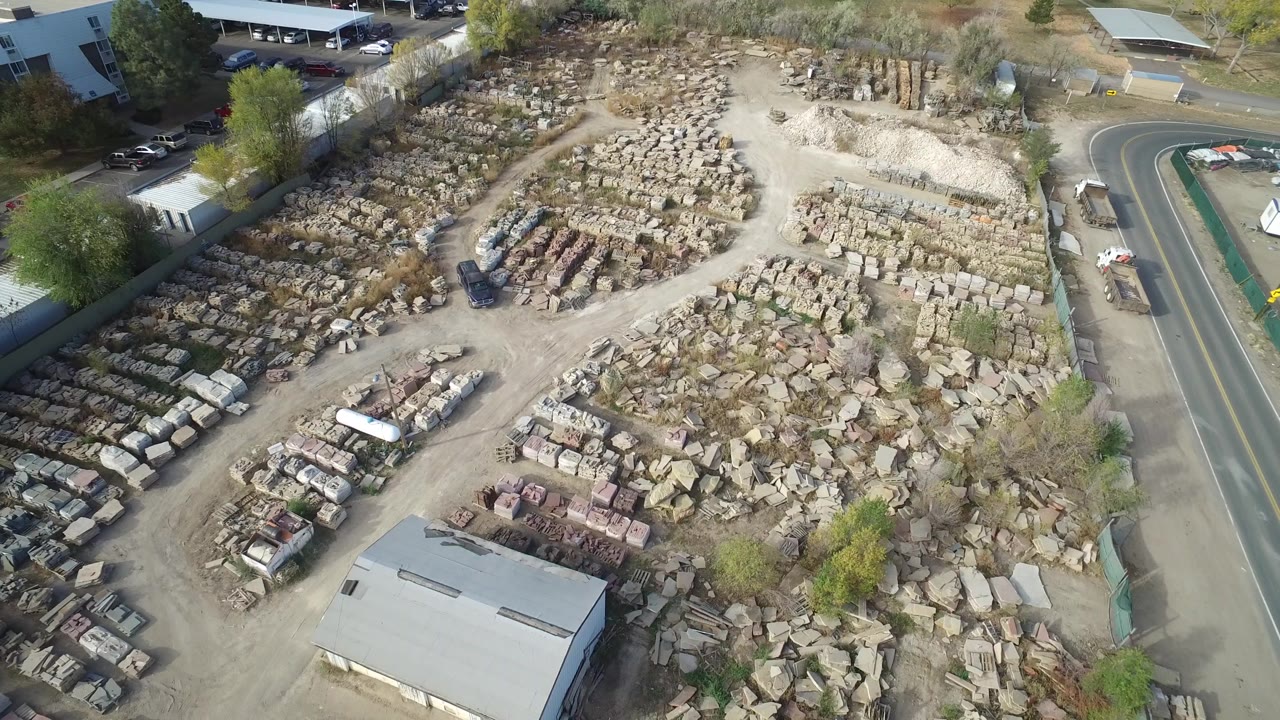 High aerial frame over a stone and materials yard in Hidden Lake, Colorado, with stacked rock rows, a low warehouse at the bottom, a paved road on the right, and no visible people.