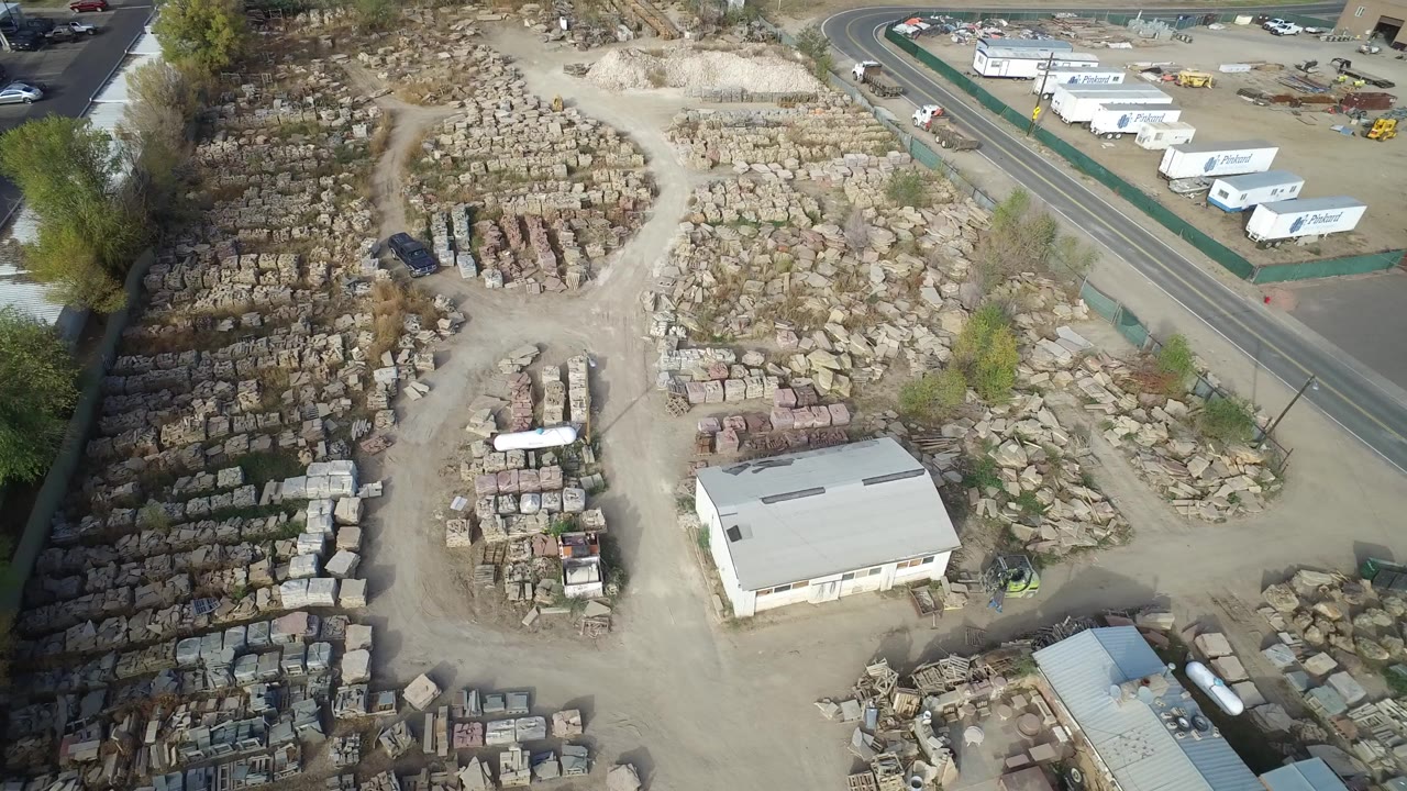 Aerial frame over Hidden Lake, Colorado showing rows of stone blocks in a dusty yard, low roofed buildings, parked vehicles near the road, and no visible people.
