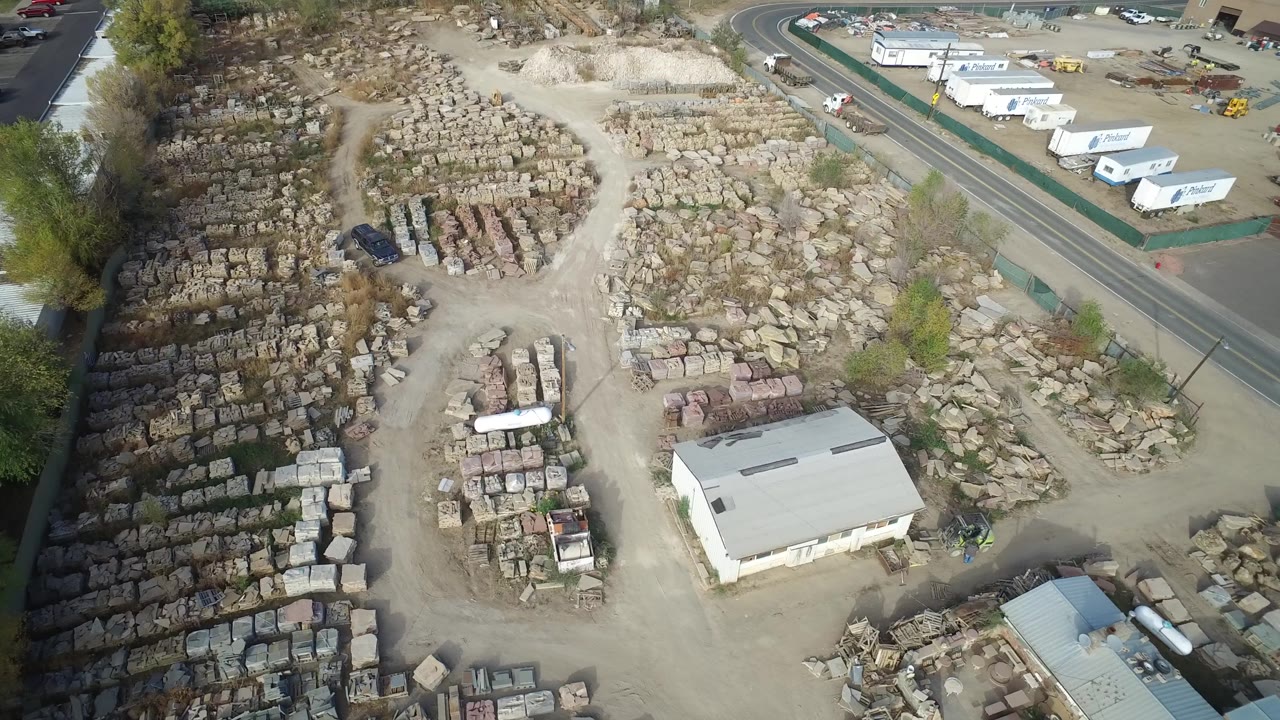 Drone-height view of a storage yard with stone piles, low buildings, parked cars, and trailers beside a road.