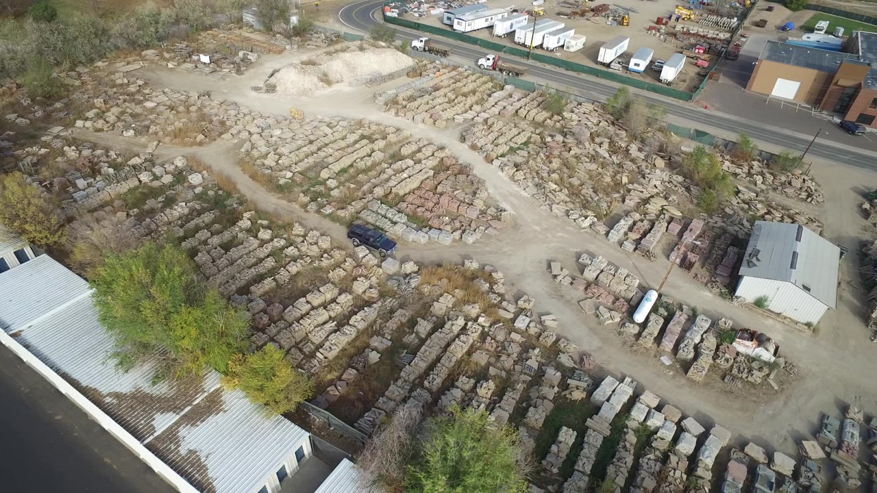 Aerial frame in Hidden Lake, Colorado showing repeating rows of stone blocks, a pale dirt yard, a roadside industrial strip, trees, parked vehicles, and no visible people.