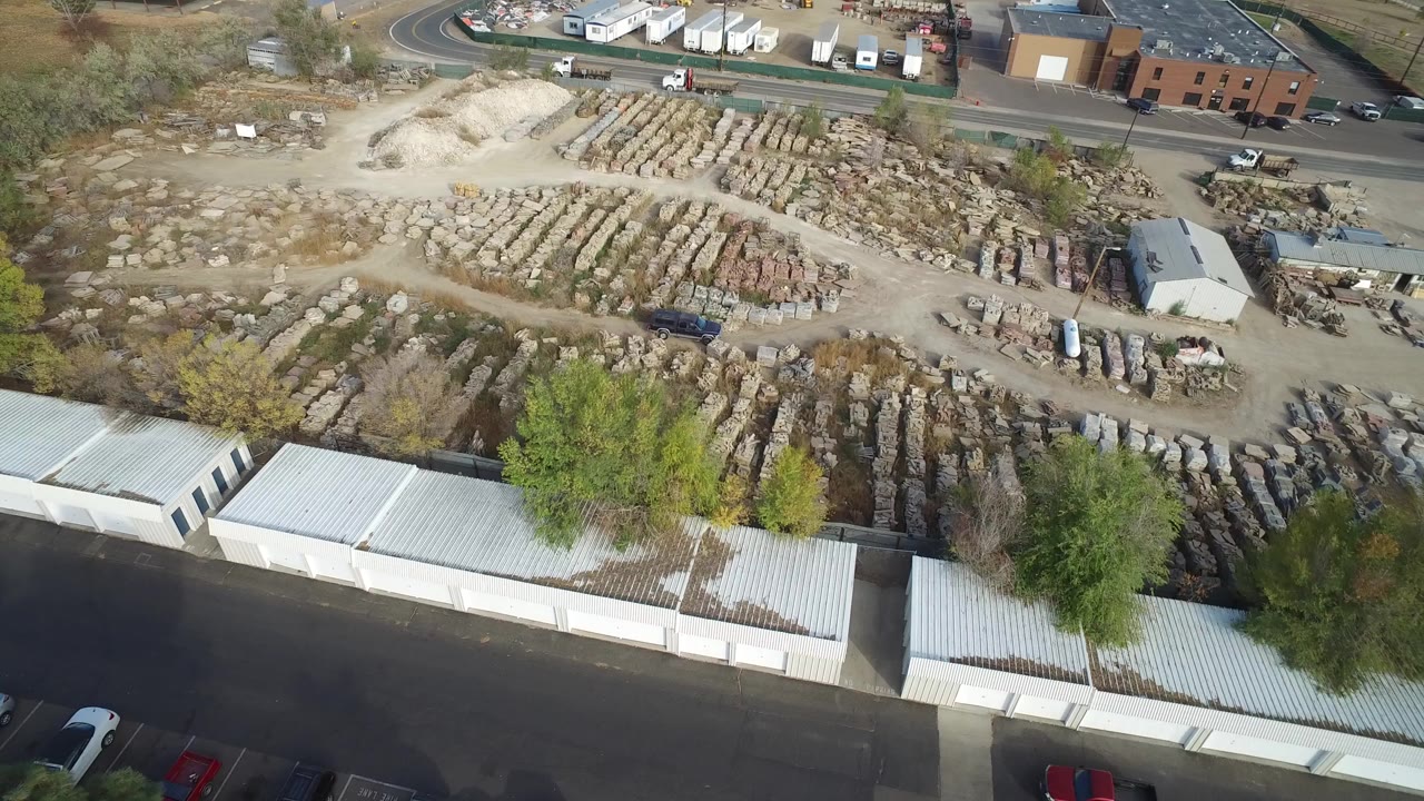 High drone view in Hidden Lake, Colorado showing rows of rocks and debris behind white storage buildings, with a road and low commercial buildings beyond and no visible people.