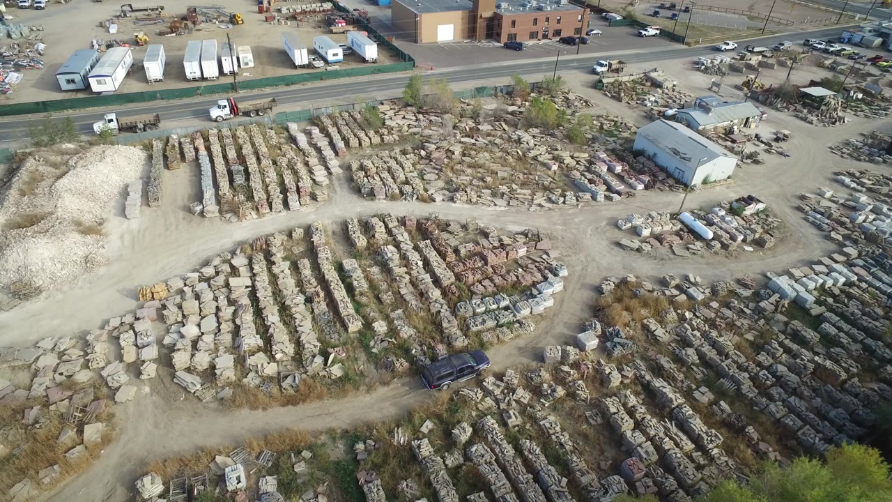 High aerial frame in Hidden Lake, Colorado, with long rows of stored material across the yard, a pale gravel mound at left, trailers and trucks near the road, and no visible people.