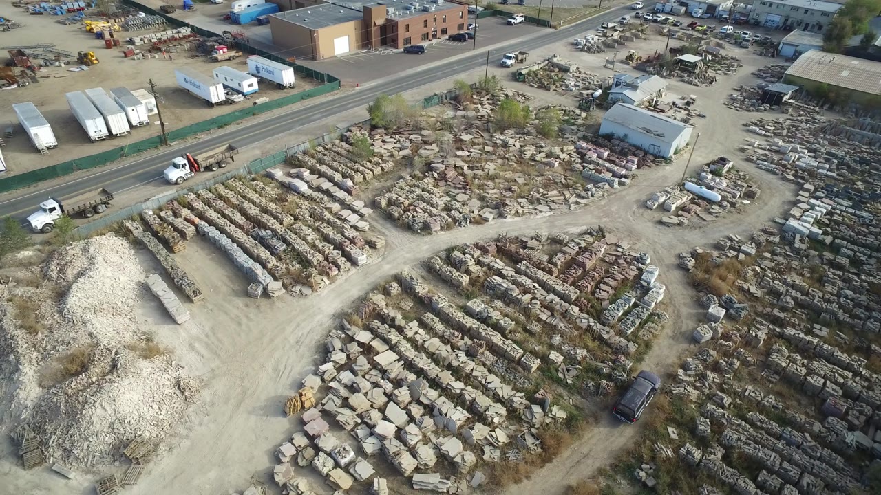 Aerial frame in Hidden Lake, Colorado with the storage yard centered, pale gravel in the lower left, parked trucks near the road, warehouse buildings beyond, and no visible people.