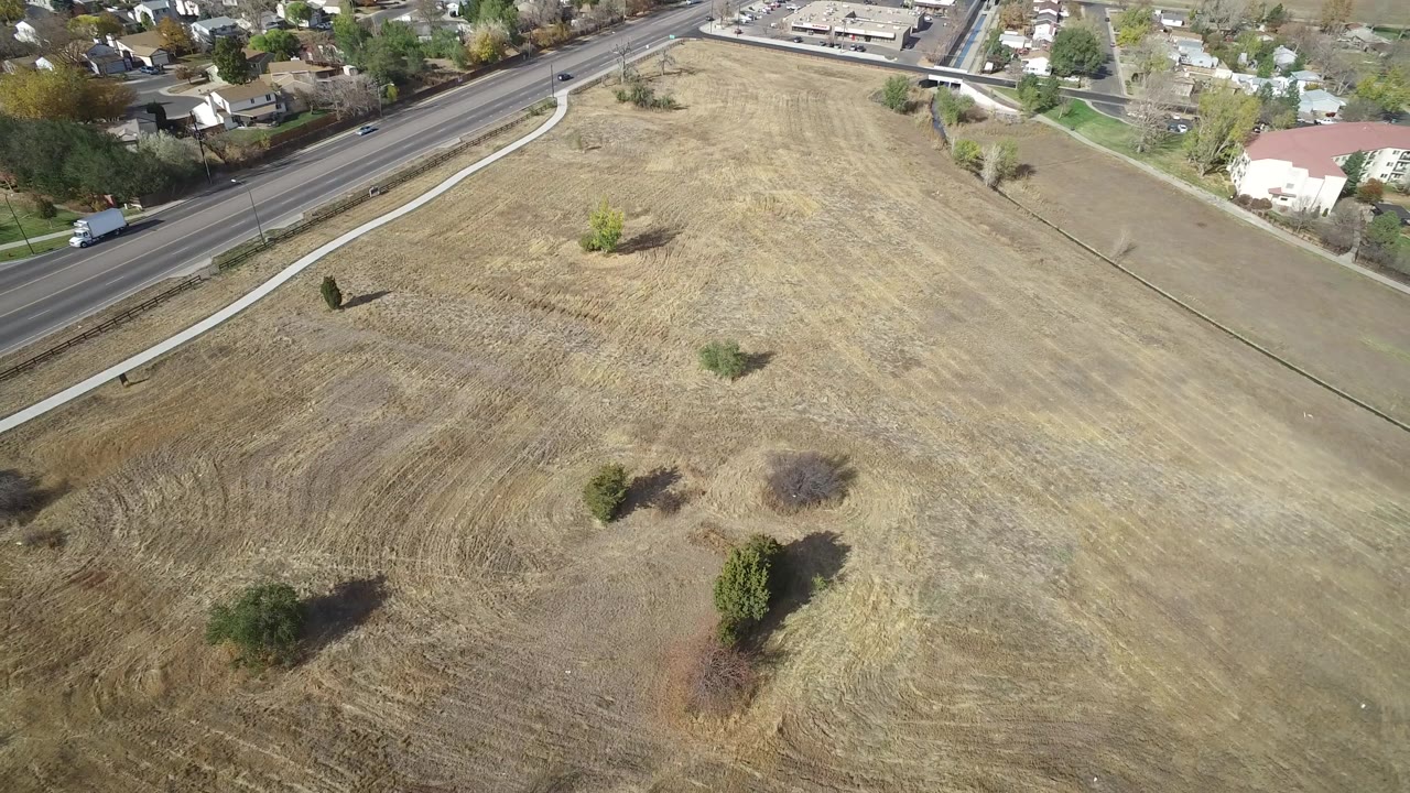 Aerial frame of the Hidden Lake, Colorado field showing tan grass patterns, small trees, a paved road with vehicles, and houses in the background with no visible people.