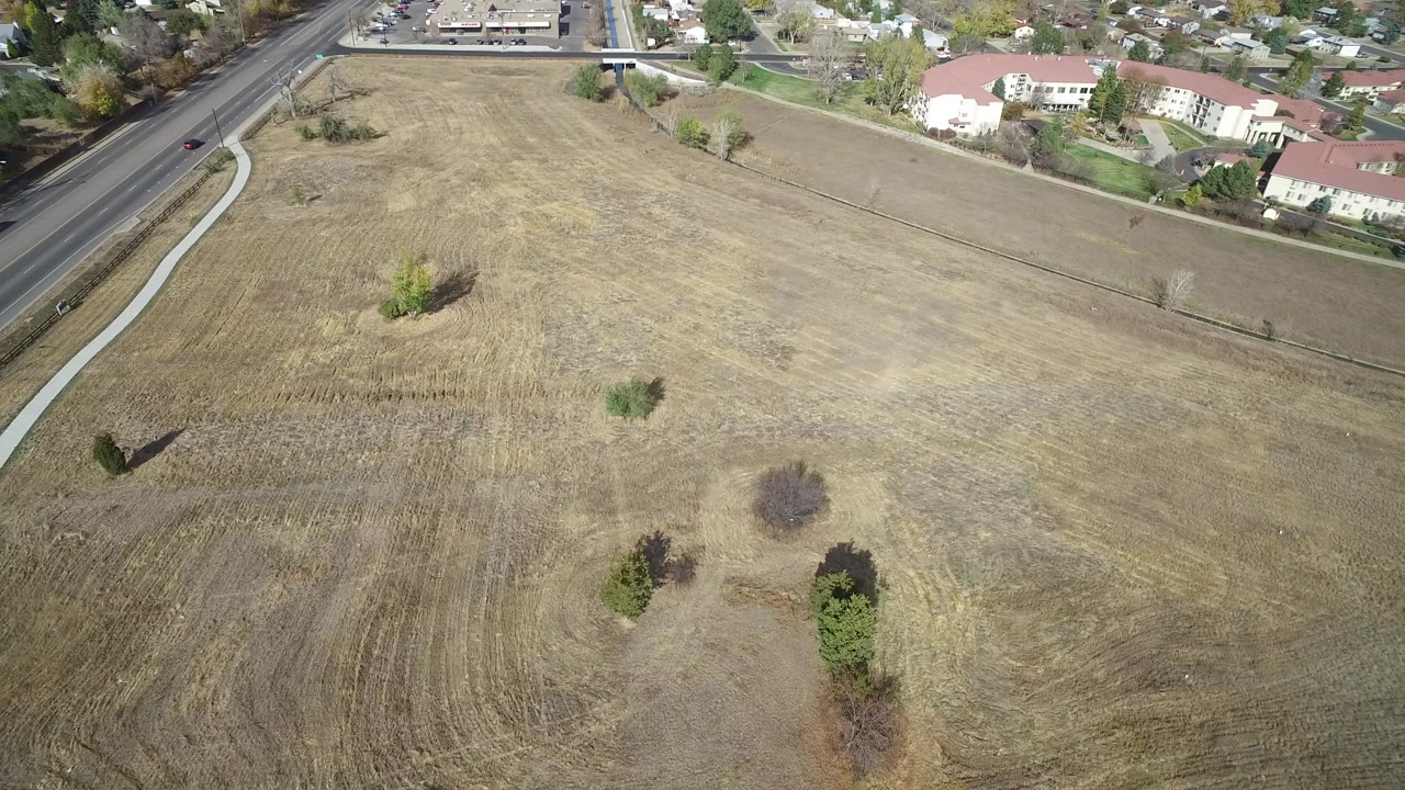 Opening aerial frame in Hidden Lake, Colorado, looking over a tan open field with scattered trees, a road at left, light-colored homes near the top, and no visible people.