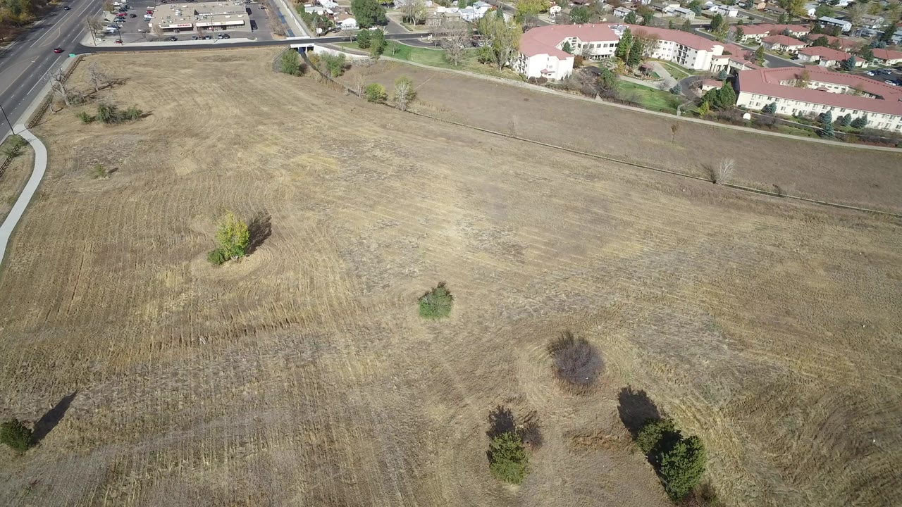 Aerial view over a dry open field with scattered trees and houses beyond.