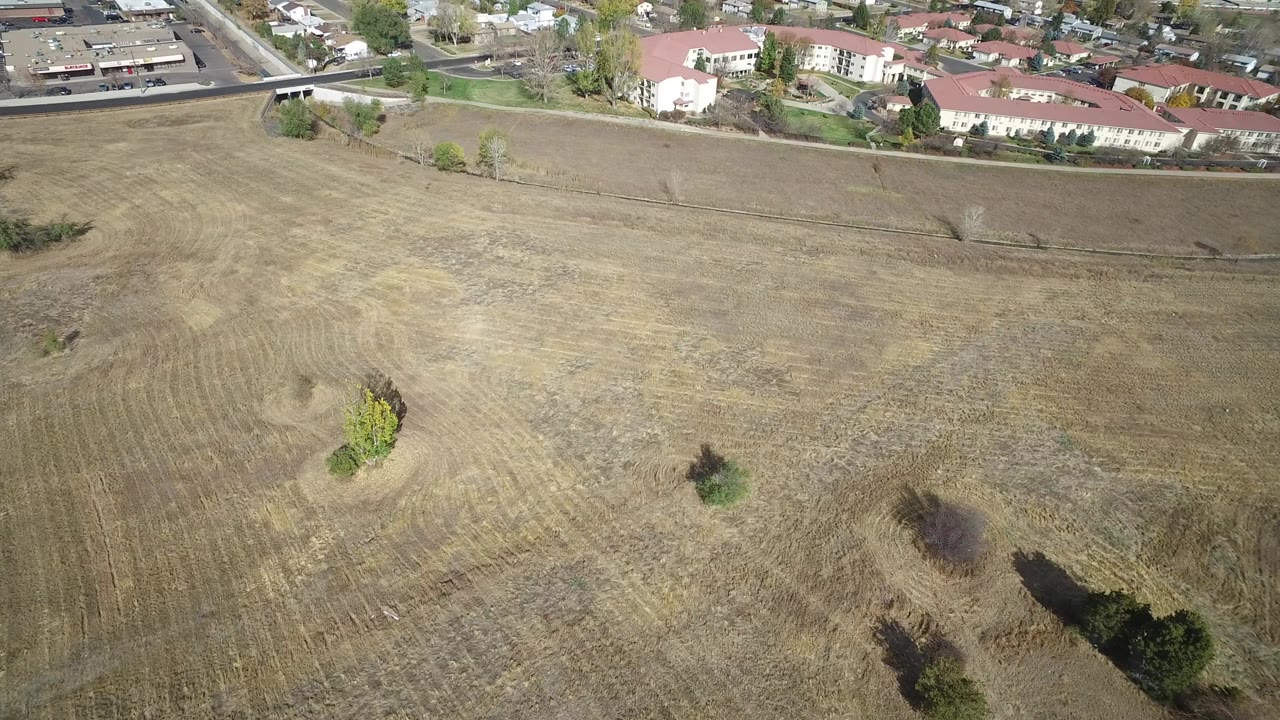 High aerial frame over a dry grassy lot in Hidden Lake, Colorado, with residential buildings and roads along the upper edge and no visible people.