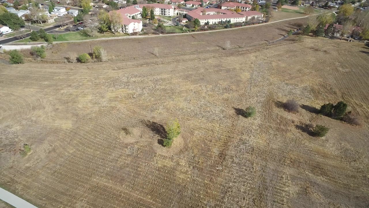 Opening aerial frame near Hidden Lake, Colorado showing a broad dry field, scattered trees, residential buildings at the top edge, and no visible people.