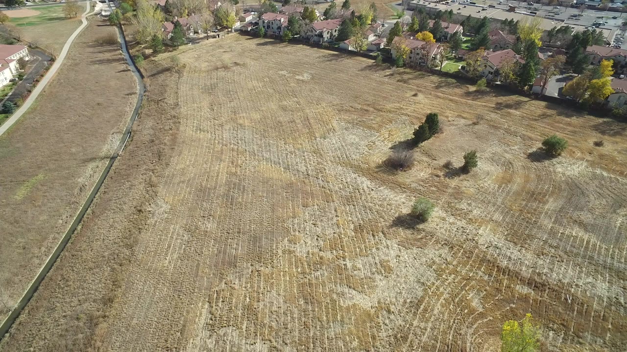Aerial frame over a dry field in Hidden Lake, Colorado, with pale crop rows, scattered trees, houses along the upper edge, and no visible people.