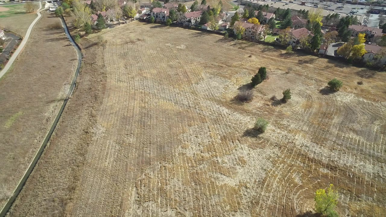 Wide aerial over tan field rows with homes beyond.