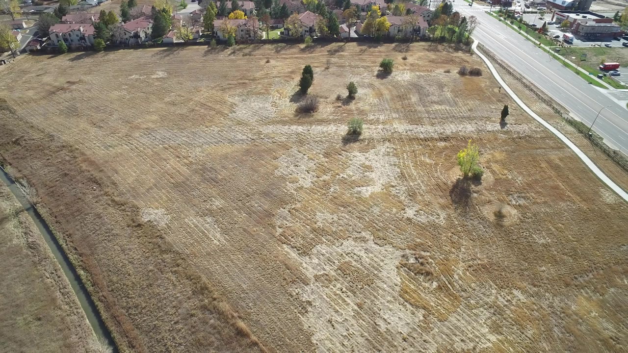 Aerial view continues across Hidden Lake, Colorado farmland, with a tan stubbled field, several small trees, a road at the right, and houses beyond it, with no visible people.