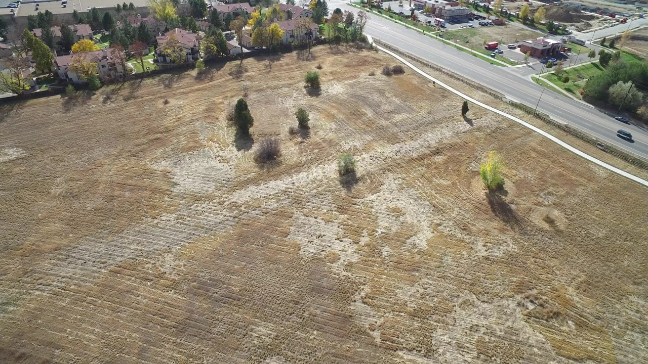 High aerial view over dry undeveloped land in Hidden Lake, Colorado, with a road, sidewalk, scattered trees, nearby houses, and no visible people.