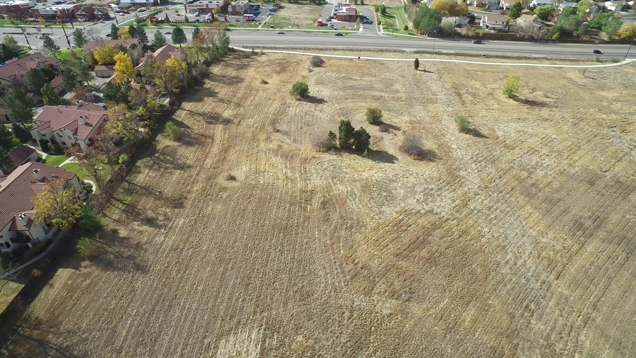 Aerial view in Hidden Lake, Colorado with the field dominating the frame, several homes tucked along the left border, small trees casting shadows, and no visible people.
