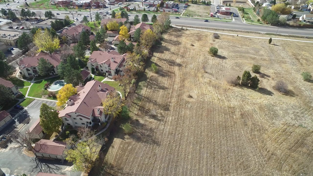 Aerial frame in Hidden Lake, Colorado with a dry field taking the right side, clustered red-roofed buildings at left, fall-colored trees, parked cars, and no visible people.