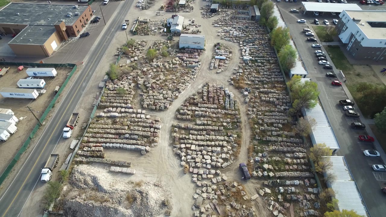 Mid-clip aerial view over Hidden Lake, Colorado, showing piles and rows of stored objects in a dusty lot bordered by roads, buildings, trailers, parked cars, and no visible people.