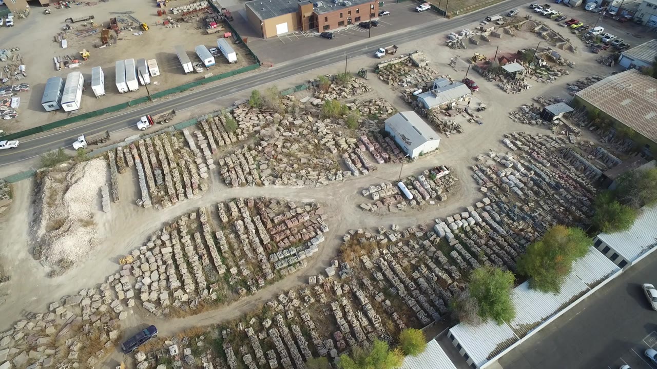 Aerial frame over Hidden Lake, Colorado with pale dirt ground, organized rows of materials, a curved road, parked buses or trailers, scattered vehicles, trees, and no visible people.