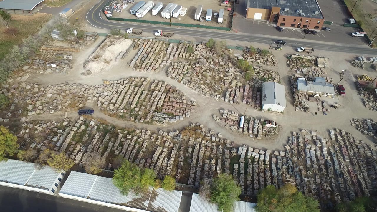 High aerial frame over Hidden Lake, Colorado, showing rows of stacked stone or masonry materials, a roadside parking area, low buildings, trees, and no visible people.