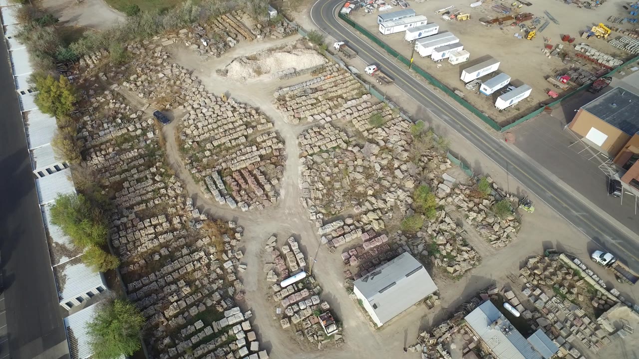 Overhead view of a dusty yard with stacked materials, sheds, trailers, road, and parked vehicles.