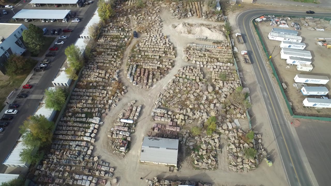 High aerial view over a Hidden Lake, Colorado roadside storage yard with rows of stone material, a small shed, parked vehicles, adjacent buildings, trees, and no visible people.