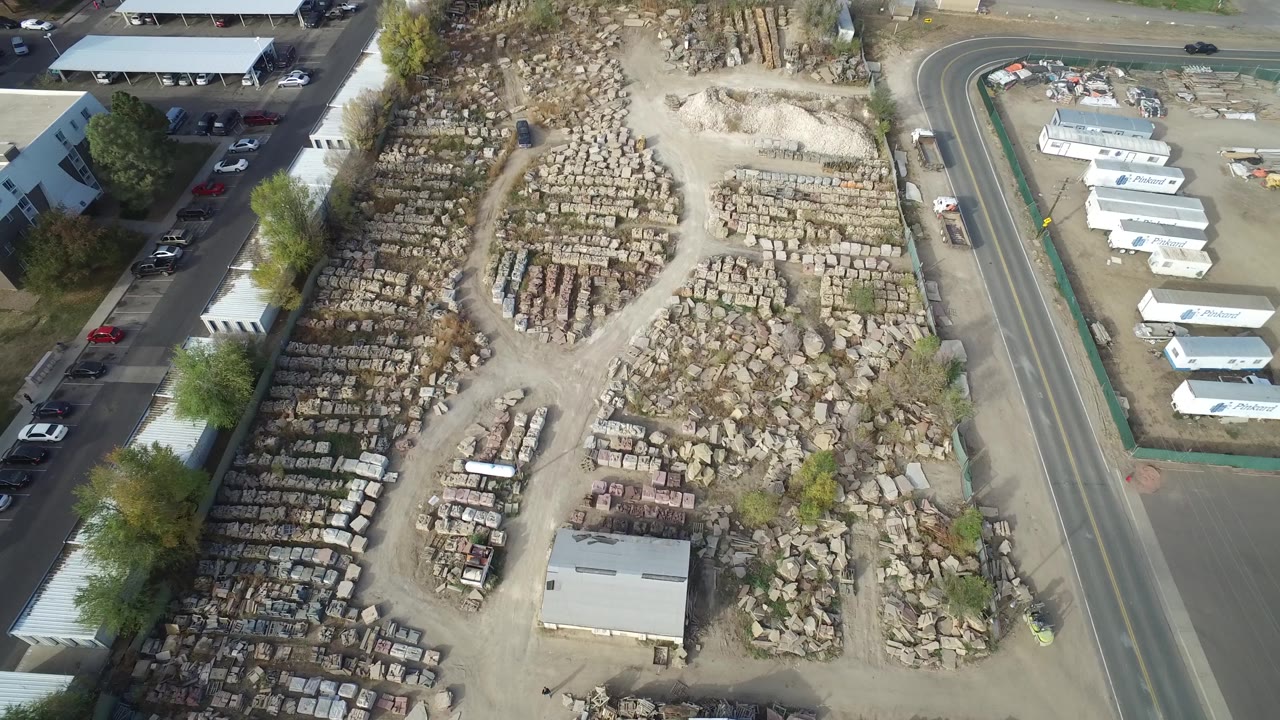 Aerial frame in Hidden Lake, Colorado showing the storage yard more centered, with a curved road to the right, white trailers beyond it, buildings at the edge, and no visible people.