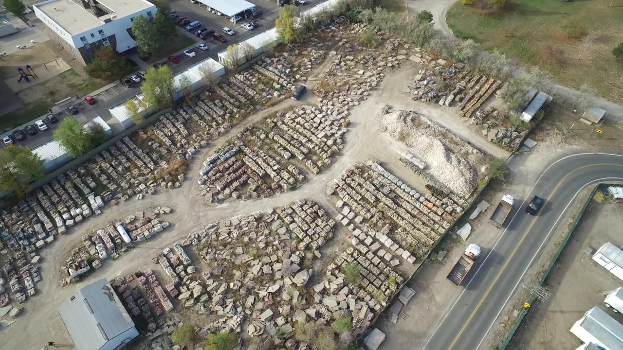High aerial view over a Hidden Lake, Colorado industrial yard with rows of stone or construction material, surrounding dirt lanes, nearby buildings, parked cars, and no visible people.