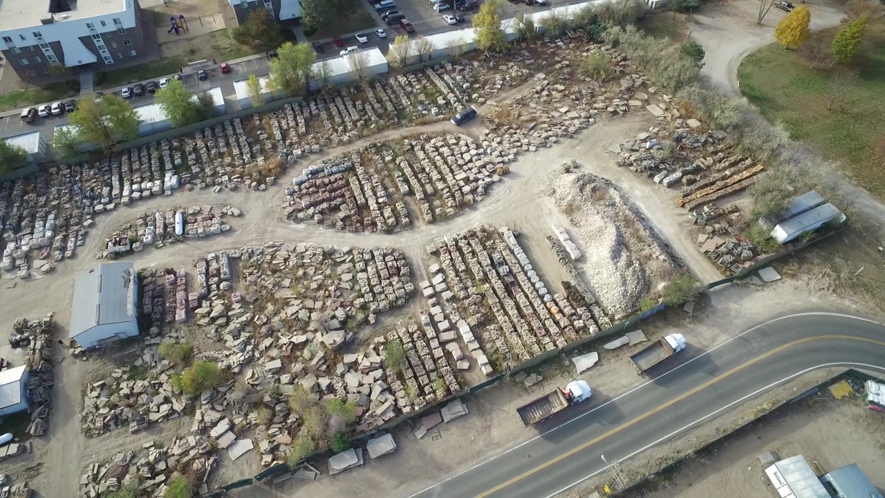 High aerial view in Hidden Lake, Colorado showing rows of wrecked cars, stone piles, dirt lanes, nearby buildings, and a curved road with no visible people.