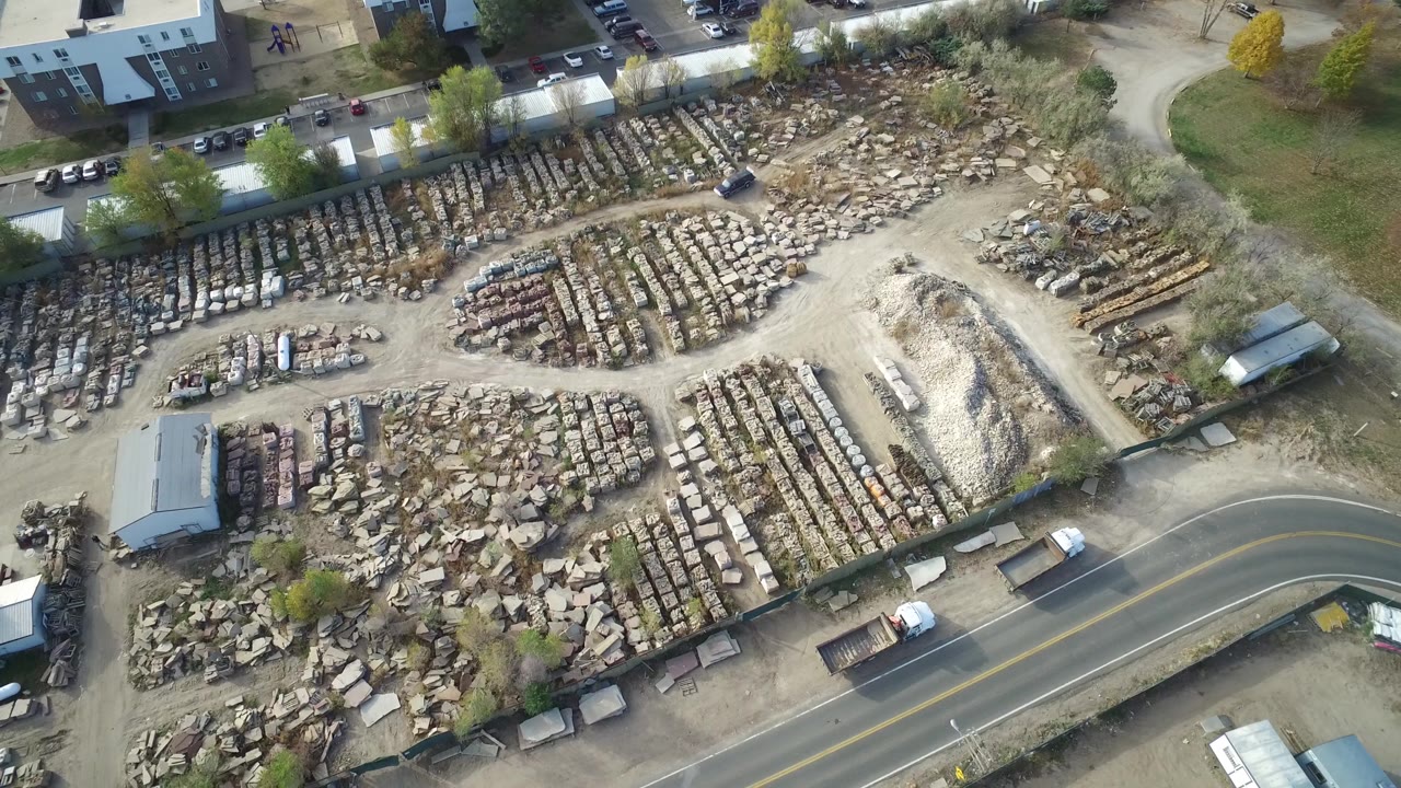 Drone-height frame of the Hidden Lake yard with gridded rows of damaged vehicles, pale dirt tracks, scrap piles, trees, and the road along the bottom, no visible people.