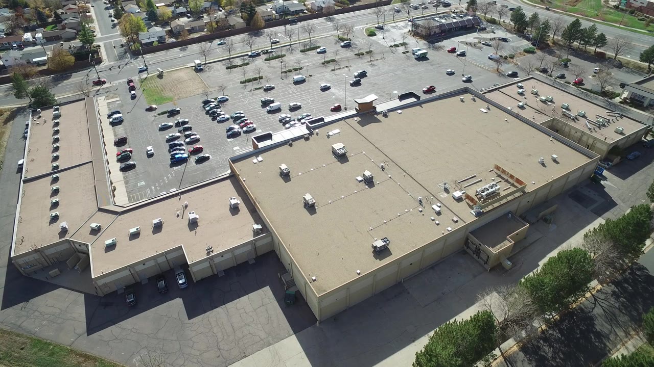 Aerial view over a tan flat-roofed building complex in Hidden Lake, Colorado, with parking lots, roads, trees, surrounding homes, and no visible people.