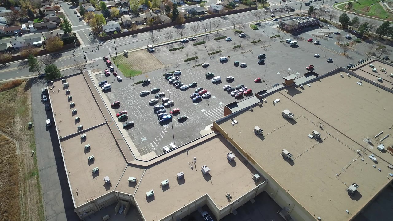 High aerial frame over Hidden Lake, Colorado showing the tan L-shaped roof in the foreground, a broad parking lot with scattered cars, streets and houses beyond, and no visible people.